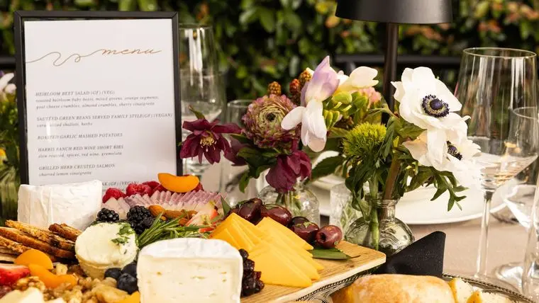 Charcuterie grazing board on a Handheld Catering event table with a printed dinner menu, fresh flowers, wine glasses and bread basket