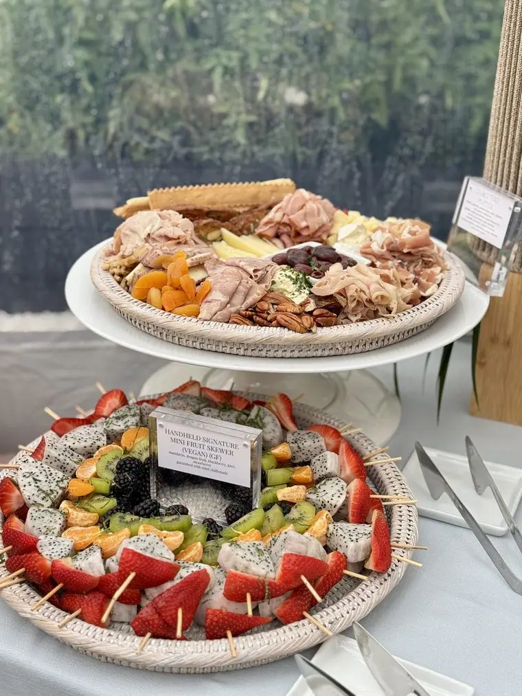 Handheld Catering baby shower charcuterie board of cured meats and cheeses above a fresh fruit platter of strawberries, kiwi and melon skewers