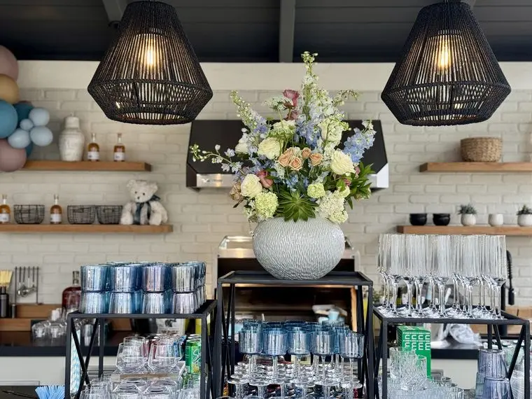 Handheld Catering baby shower bar station with polished glassware, stemware and chilled bottles beneath a large seasonal floral arrangement