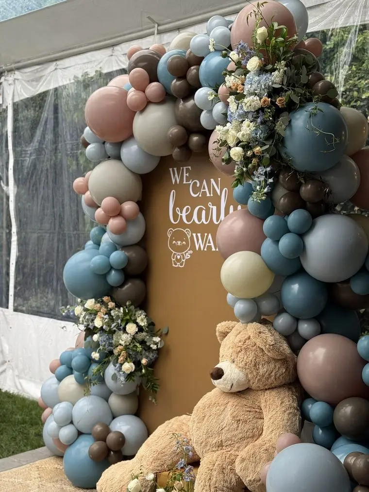 Handheld Catering baby shower balloon arch in blue, taupe and cream framing a welcome sign with a large teddy bear and dried floral accents