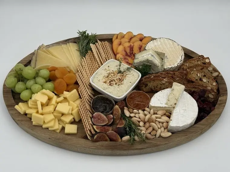 Handheld Catering artisan local cheese board with brie, manchego, cubed cheddar, hummus, peaches, figs, grapes, dried apricots, marcona almonds and crisps on a round wood platter