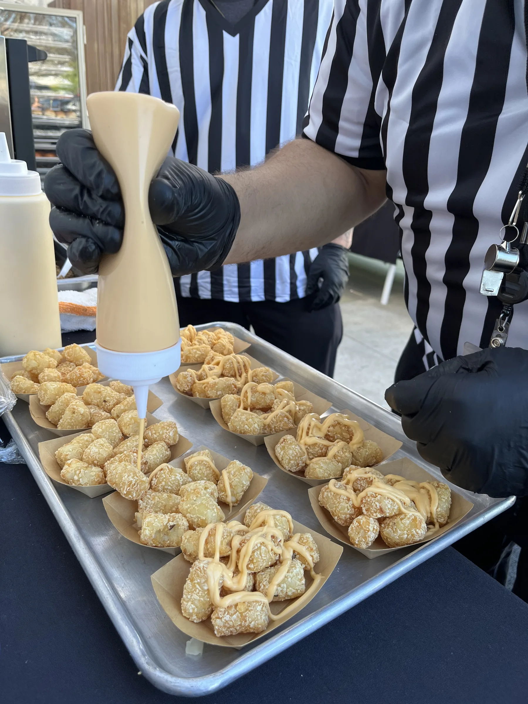 Handheld Catering referee-uniformed team member finishing fresh Tater Tot Nachos with cheese sauce during Silicon Valley corporate March Madness event