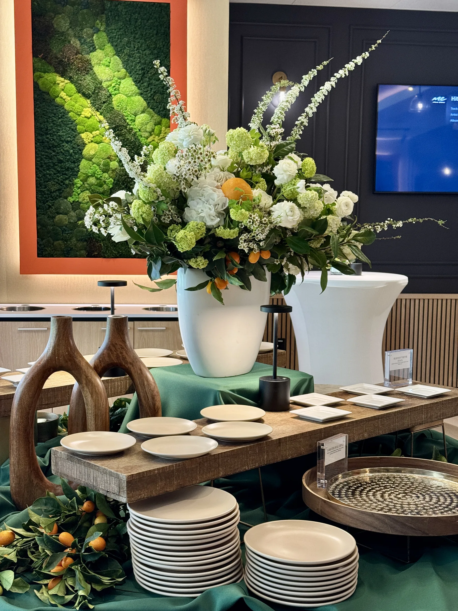 Elegantly styled catering table with large white floral arrangement, stacked plates, and green linen set for Handheld Catering's grand opening milestone celebration in Santa Clara