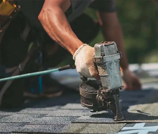 Man on roof of a home, holding a nailing gun.