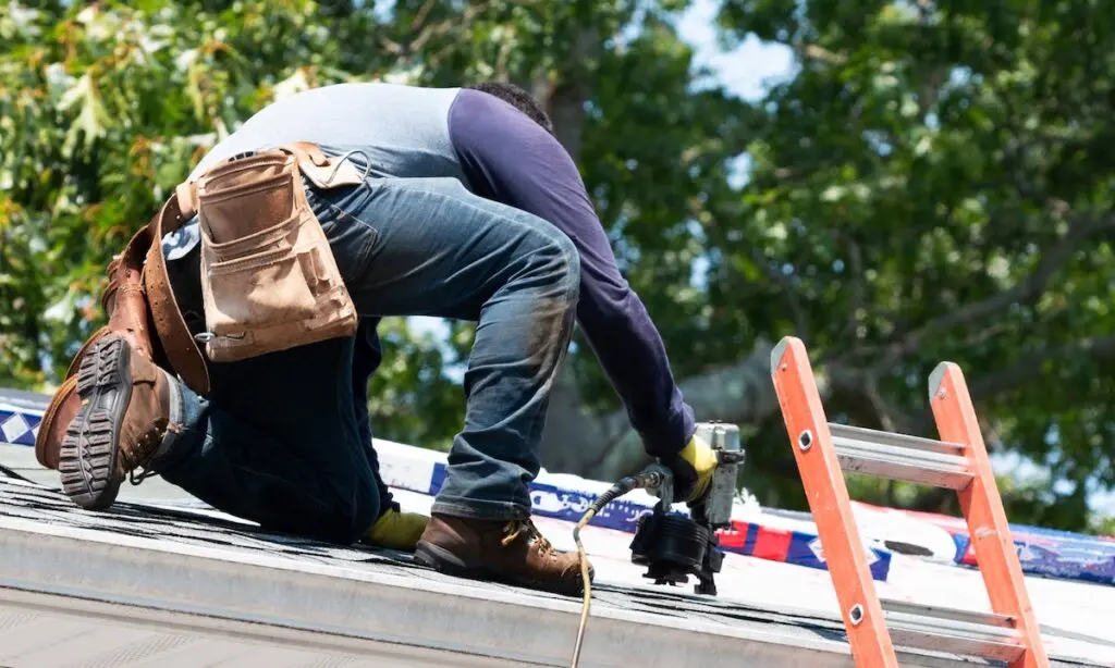 A contractor is kneeling on a residential roof using a nailgun to put new roof shingles on a house next to a ladder.