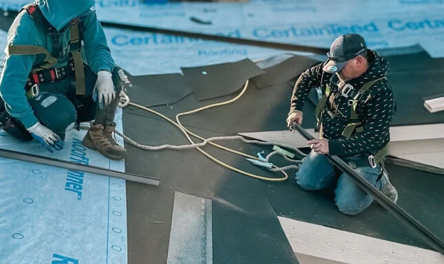 Two roofers in safety harnesses installing shingles over CertainTeed underlayment