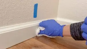 Gloved hand wipes a white baseboard with a cloth, preparing it for painting or repair. Blue tape is visible marking an area of attention along the wall.
