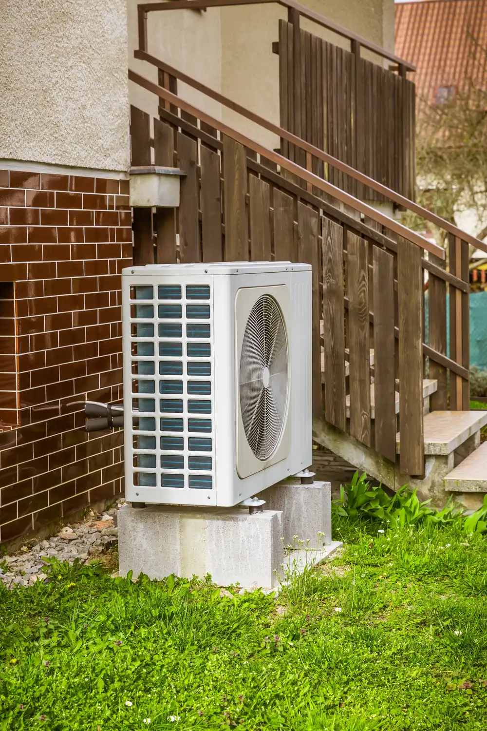 Outdoor HVAC unit on concrete blocks beside a brick wall and wooden staircase.