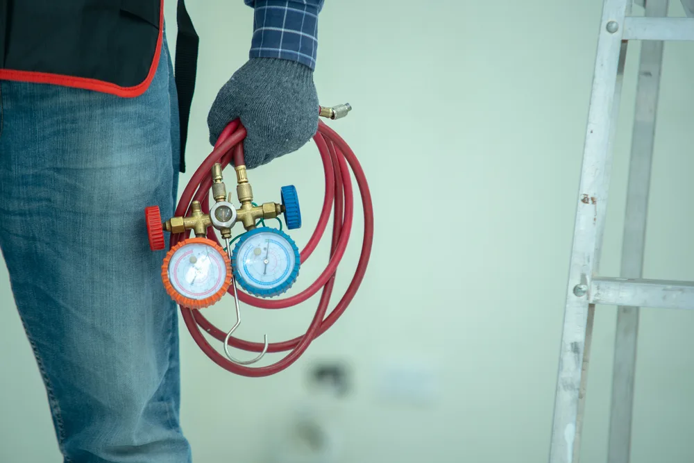 Technician holding HVAC manifold gauges next to a ladder.
