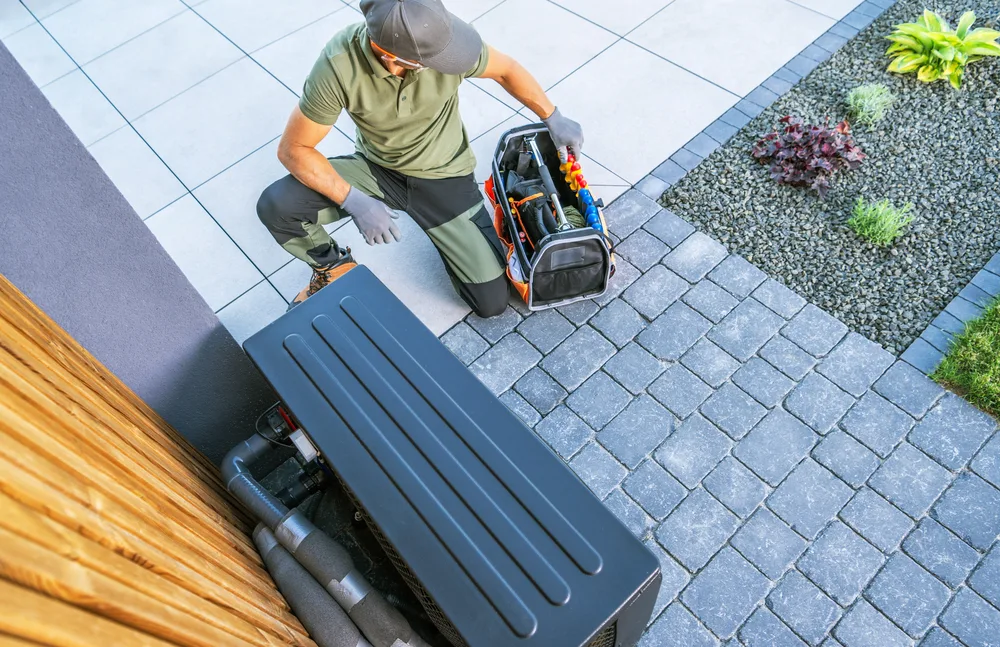 Technician kneeling beside an outdoor HVAC unit with a tool bag on a paved patio.