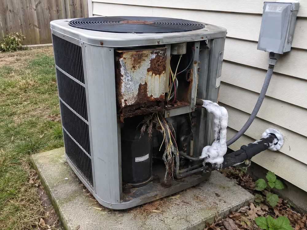 Rusty outdoor air conditioning unit with exposed wires and ice buildup on pipes.