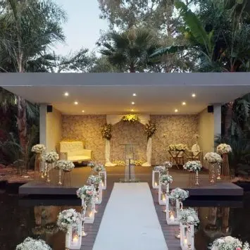 Romantic outdoor ceremony setup at The Garden Venue wedding venue, featuring a modern stage decorated with floral arrangements, a white loveseat, and soft lighting, with a flower-lined walkway over a tranquil pond surrounded by lush greenery.