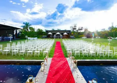 Outdoor ceremony setup at The Garden Venue wedding venue, featuring rows of white chairs arranged on a manicured lawn, a striking red carpet aisle over a reflective pond, and elegant floral arrangements under a vibrant blue sky.