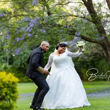 Bride and groom joyfully dance together on a lush green lawn beneath blooming jacaranda trees at The Garden Venue wedding venue, with the bride holding a bouquet and both smiling brightly.