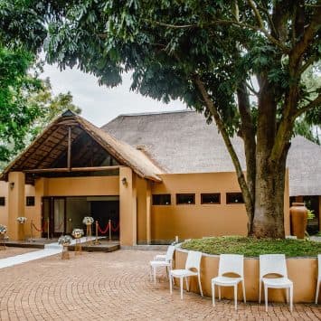 Exterior view of a thatched-roof building at The Garden Venue wedding venue, featuring a circular courtyard with white chairs, a tree at the center, floral arrangements, and a white aisle runner leading to the entrance.