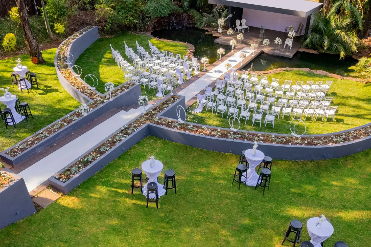 Forest Wedding Page Trio 2 Outdoor wedding venue featuring a wooden walkway over a pond leading to a floral arch, surrounded by white chairs arranged in a semi-circle on a lush green lawn under tall trees.