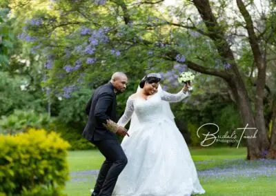 Bride and groom joyfully dance together on a lush green lawn beneath blooming jacaranda trees at The Garden Venue wedding venue, with the bride holding a bouquet and both smiling brightly.