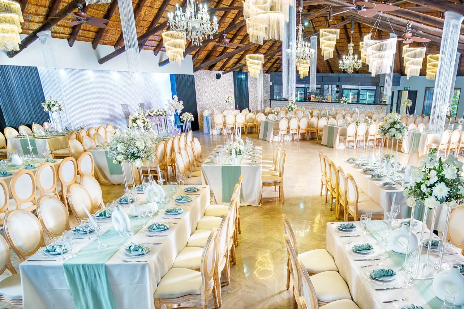 Wedding Page Trio Elegant wedding venue reception setup with round tables decorated with gold accents, floral centerpieces in pink, white, and purple tones, gold cutlery, and gold chairs, all arranged in a room with large windows overlooking greenery.
