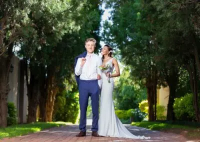Bride and groom pose playfully on a tree-lined path at The Garden Venue wedding venue, smiling brightly.