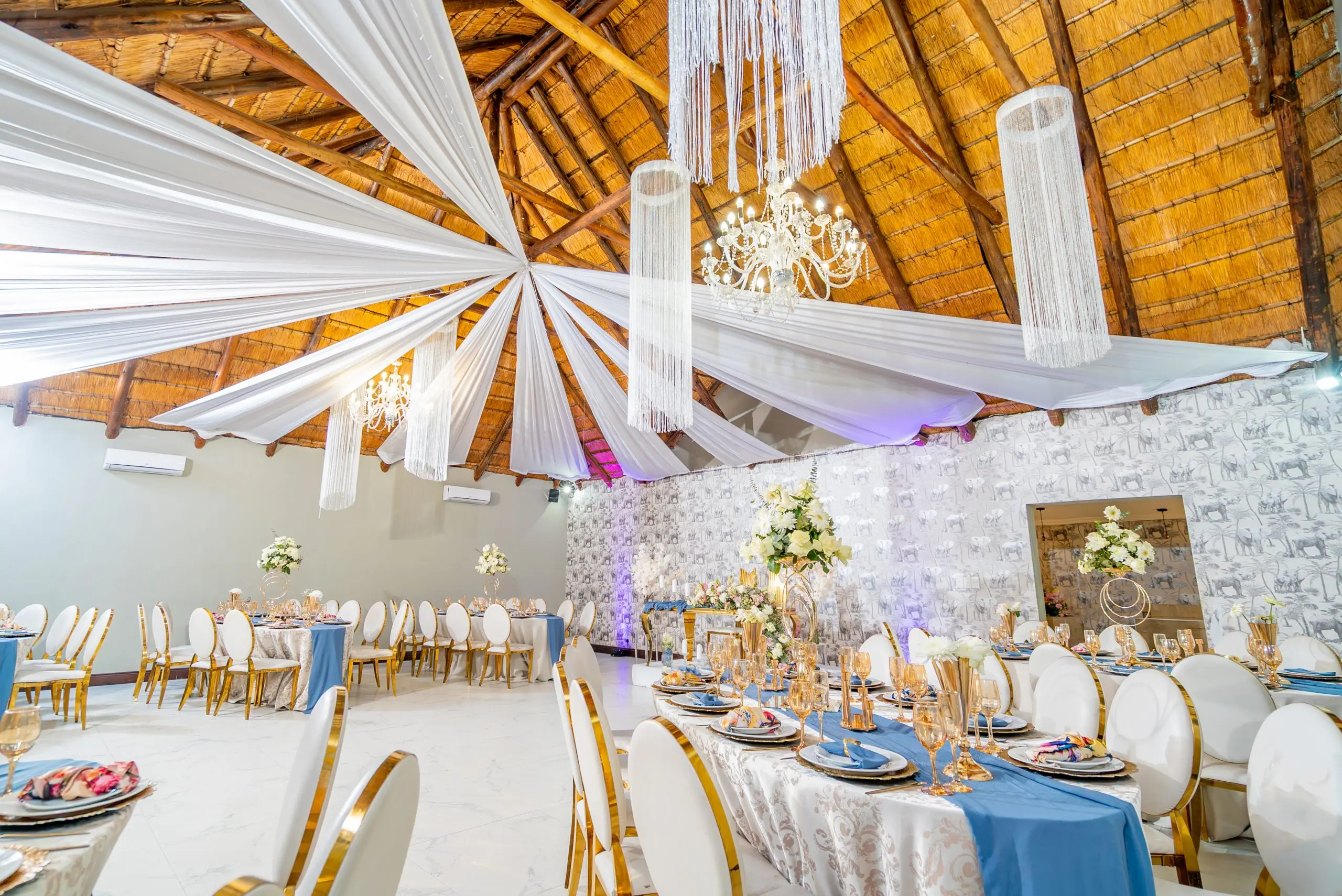Elephant-Enhanced-NR (1) Indoor wedding venue reception area with elegant white and gold chairs, long tables decorated with green runners, floral centerpieces, and formal place settings, set beside large windows overlooking lush greenery.