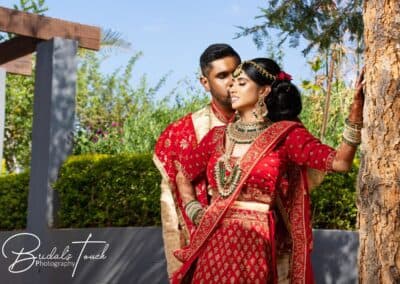 Bride and groom in traditional Indian attire pose affectionately under the sunlight at The Garden Venue wedding venue, with the bride leaning against a tree and lush greenery in the background.