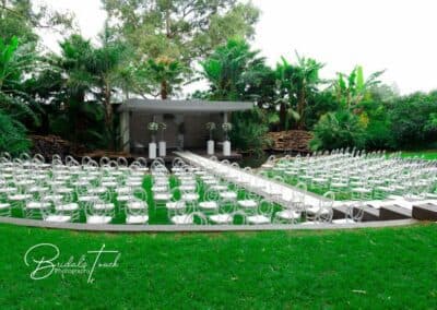 Outdoor ceremony setup at The Garden Venue wedding venue, showcasing a modern stage with white floral arrangements, surrounded by clear chairs arranged on lush green grass and a wooden walkway crossing over a tranquil water feature.