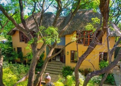 Charming wedding venue building with a thatched roof and yellow walls, surrounded by lush greenery and tall trees, creating a secluded and serene atmosphere.