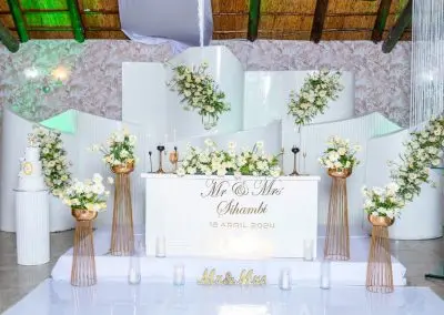 Elegant indoor wedding setup at The Garden Venue wedding venue, showcasing a modern white stage with gold accents, floral arrangements in white and yellow tones, candles, and a personalized sweetheart table for the bride and groom.