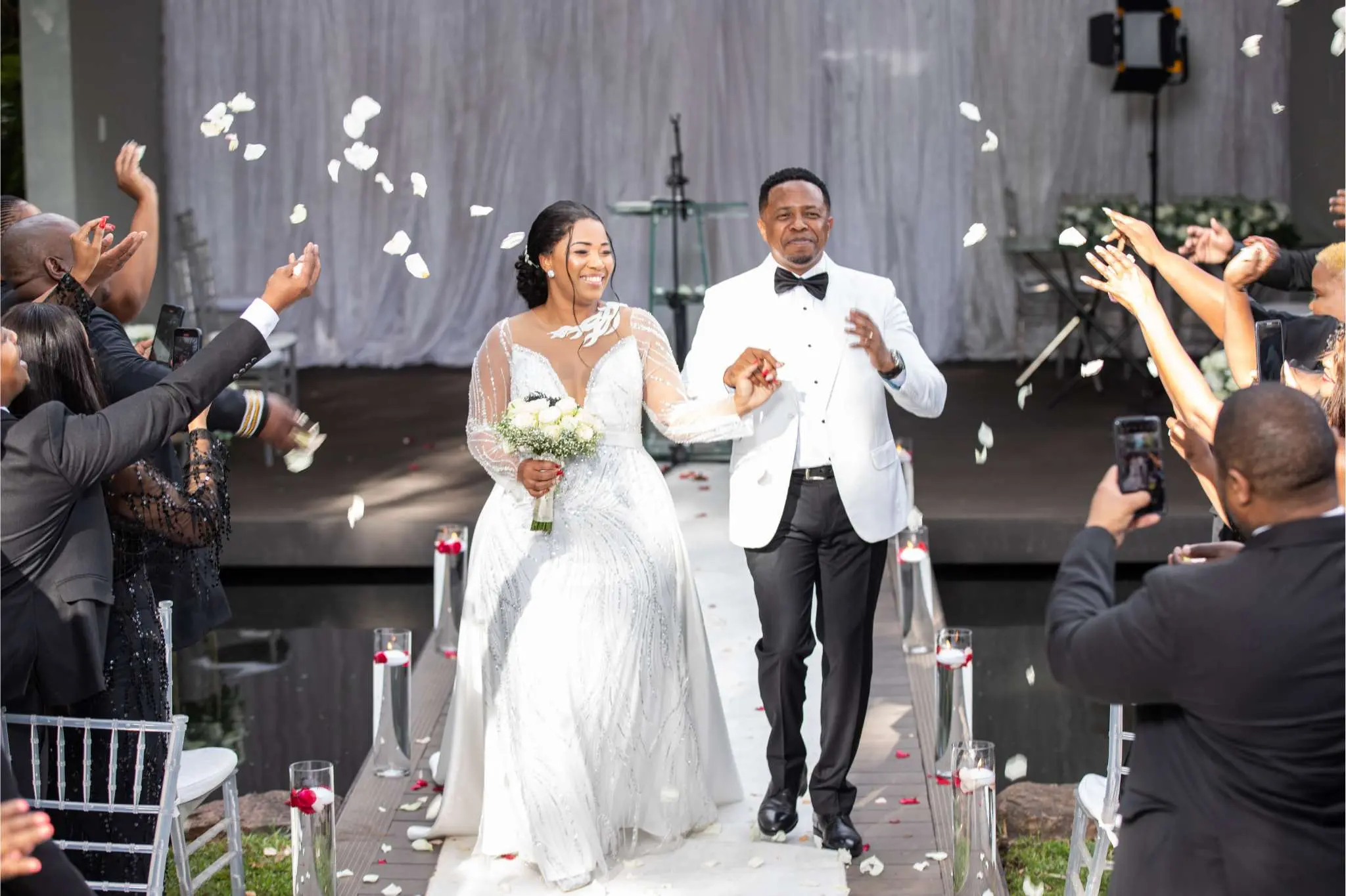 Bride and groom walk down the aisle at The Garden Venue wedding venue, smiling joyfully as guests throw flower petals in celebration, with a white-draped stage and elegant décor in the background.