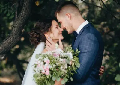 Bride and groom share an intimate moment beneath a tree at The Garden Venue wedding venue, with the groom gently holding the bride’s face and the bride smiling while holding a bouquet of pink and white flowers.