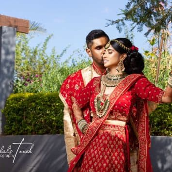 Bride and groom in traditional Indian attire pose affectionately under the sunlight at The Garden Venue wedding venue, with the bride leaning against a tree and lush greenery in the background.