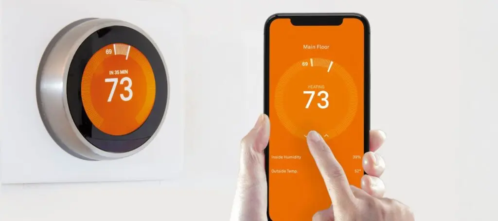 smart thermostat positioned on the wall with the phone that controls it being held in front of it being adjusted by the hands that are holding it