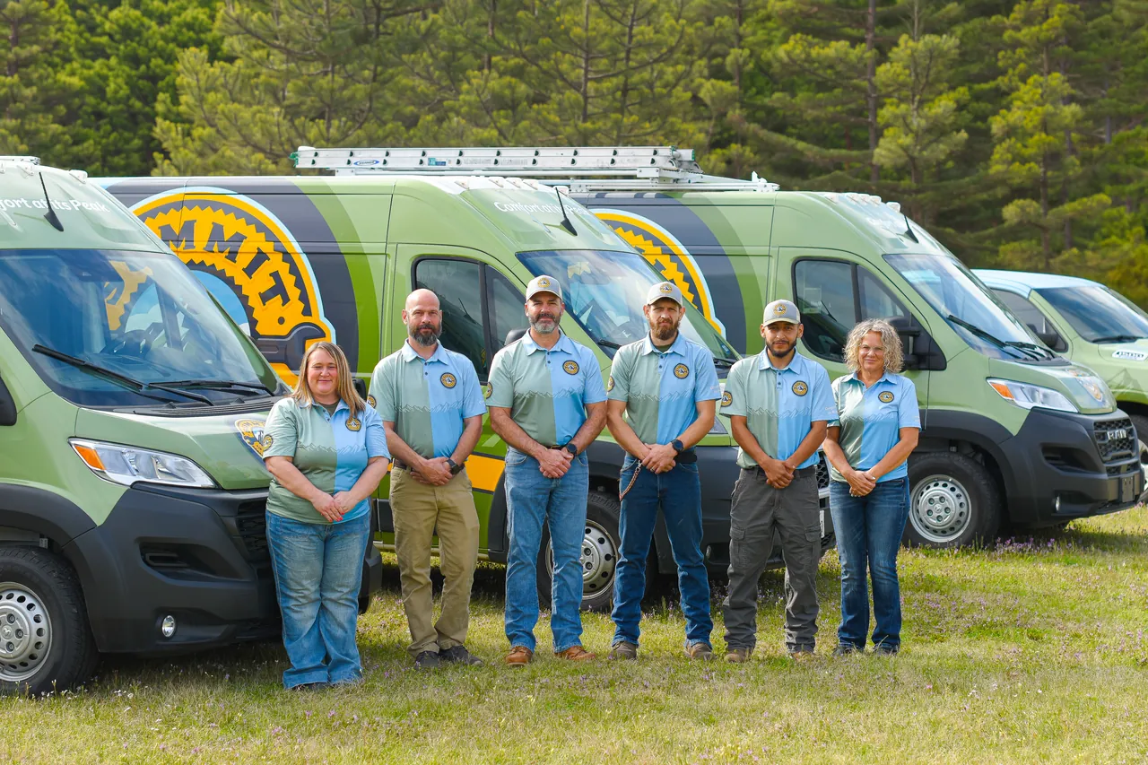 Team of six people in matching shirts standing in front of green service vans with trees in the background.