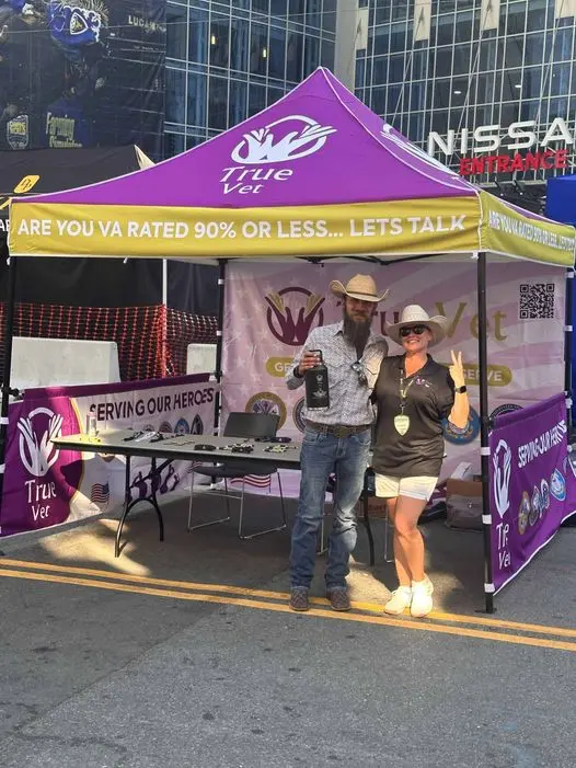 A man and woman standing under a purple True Vet tent at an outdoor event.