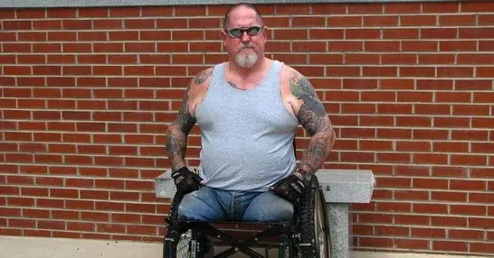 Veteran in wheelchair with tattoos standing against brick wall.