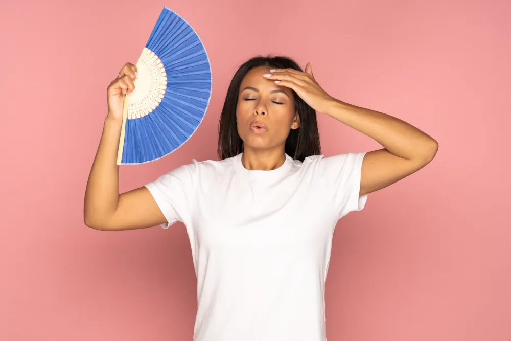 woman using paper fan suffer from heat sweating, feels sluggish isolated on pink.