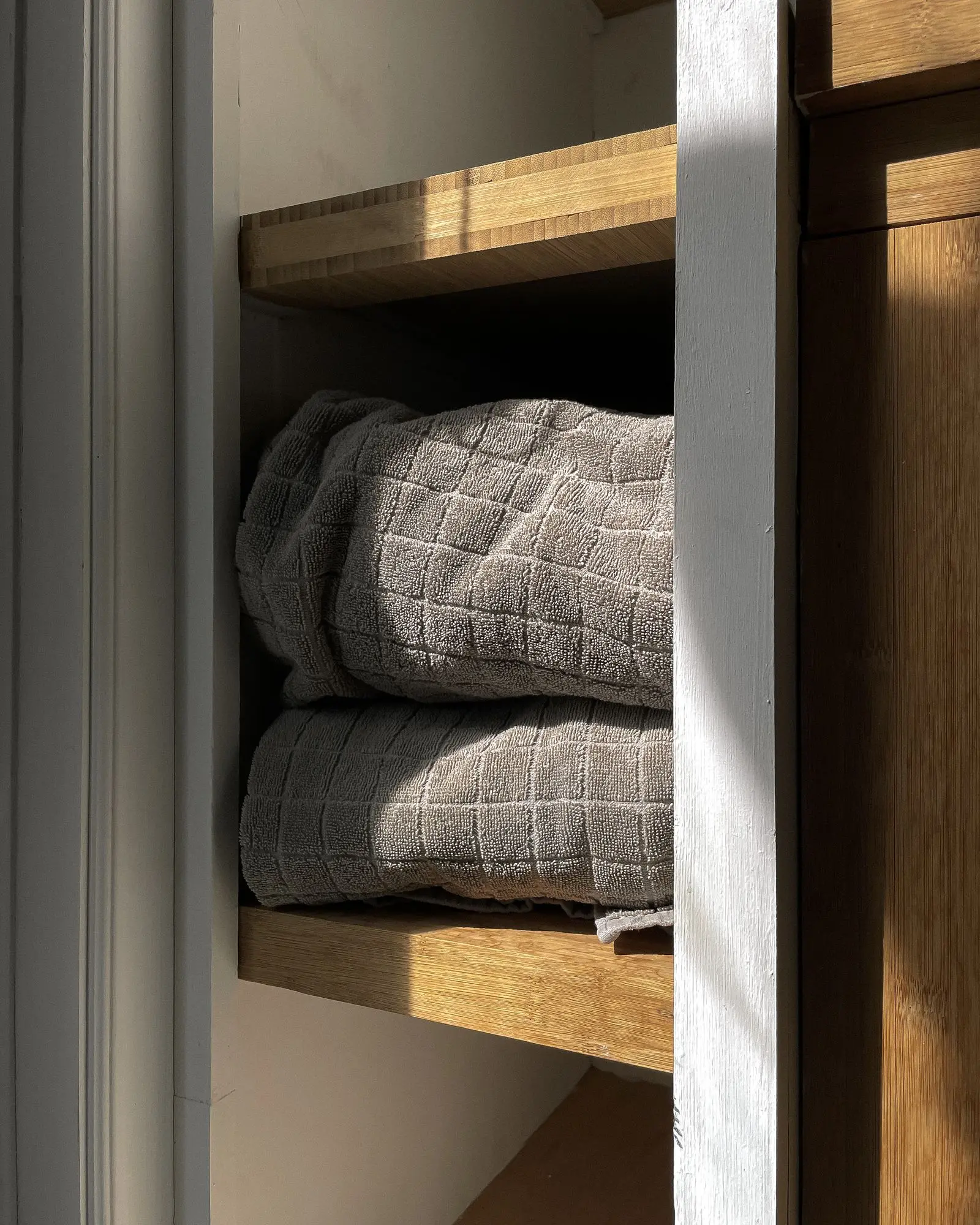 Gray textured towels folded on a wooden bathroom shelf in warm sunlight