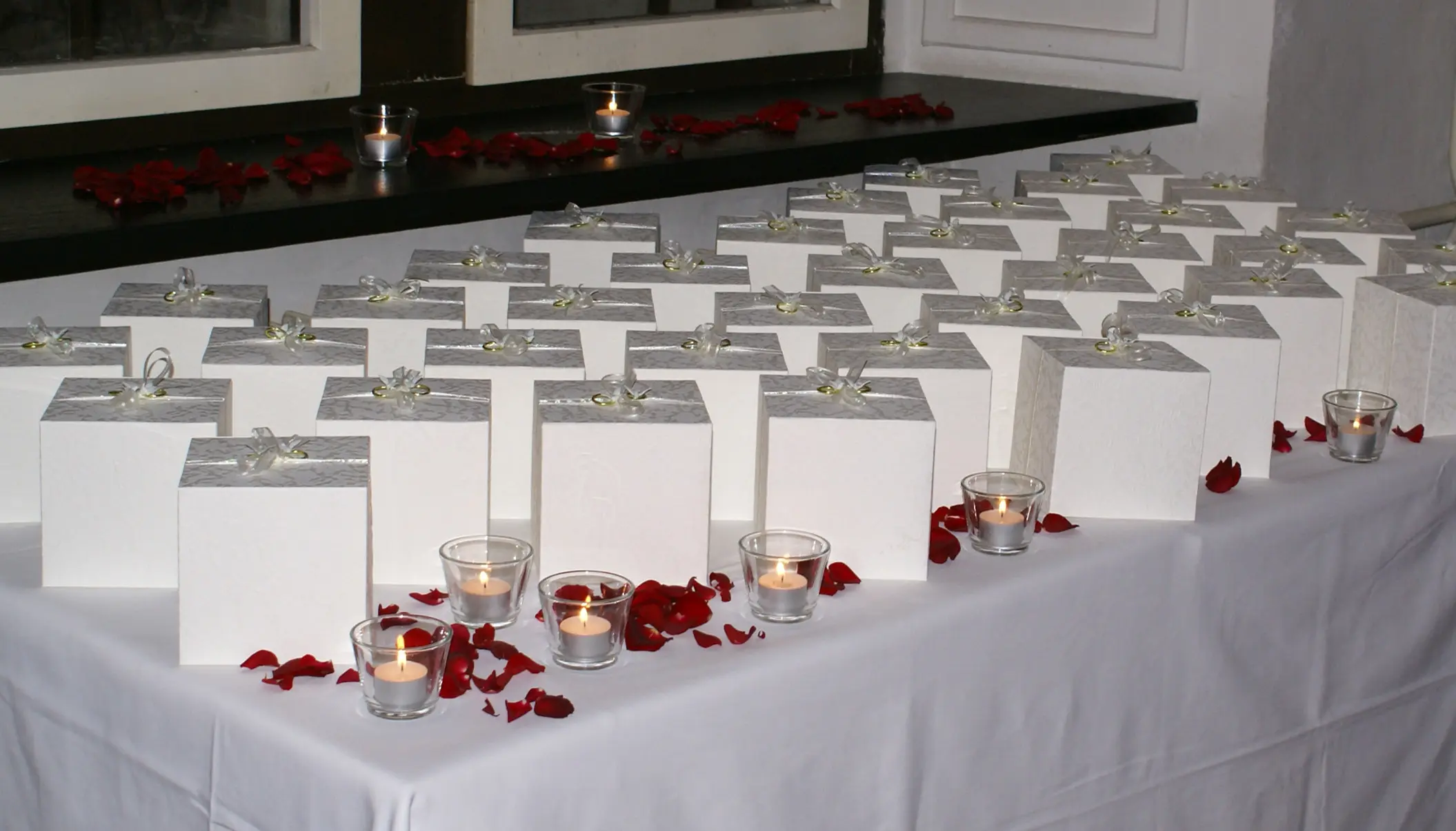 White event favor boxes arranged on a table with candles and red petals