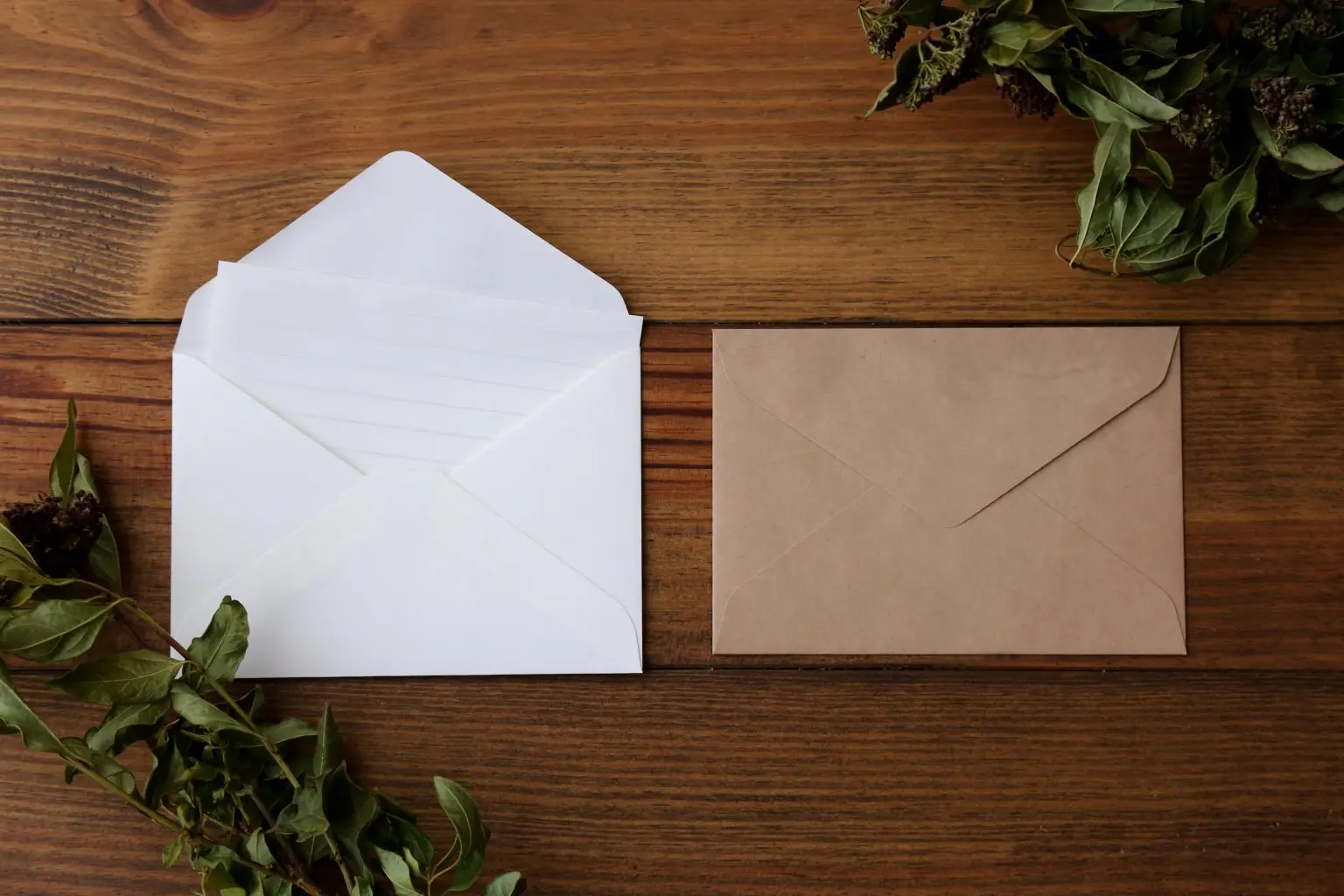 Blank white and kraft envelopes with dried foliage on a wooden table