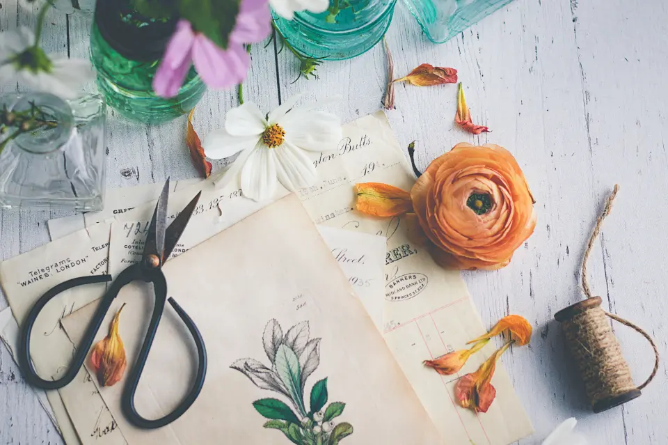 Botanical flatlay with flowers, scissors, paper, and glass jars on a white wood surface