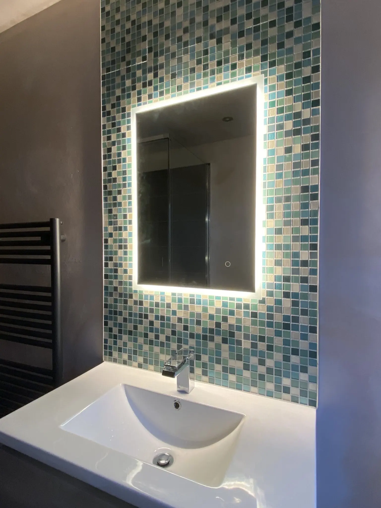Illuminated rectangular bathroom mirror above a white sink and blue mosaic tiles