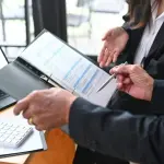 Two professionals discuss documents while a laptop and calculator are on the table.