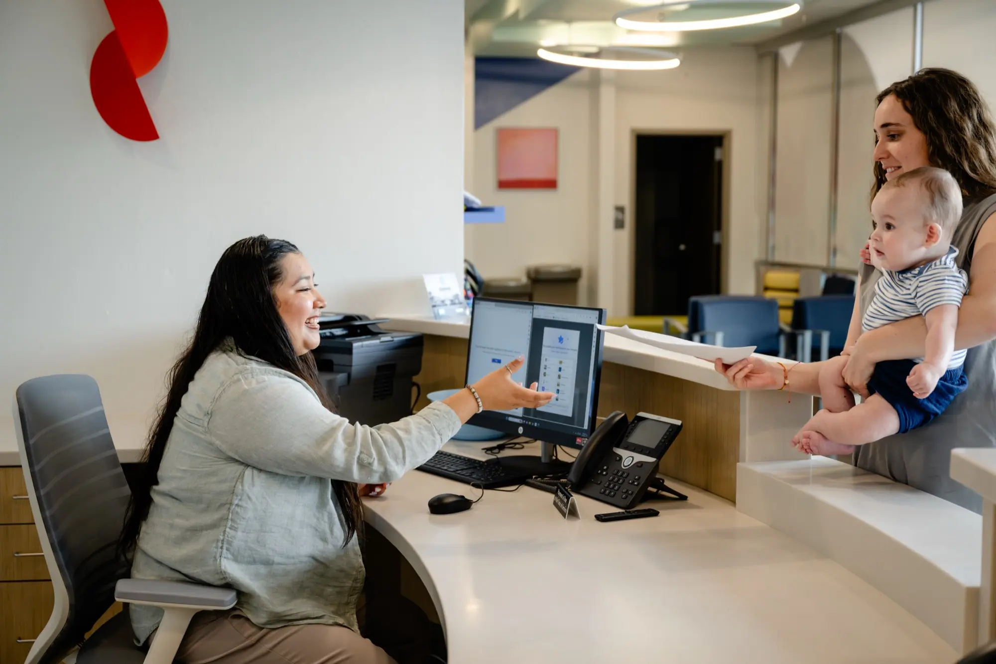 A parent holding a baby checks in with a smiling team member at the Care+ Edmond front desk