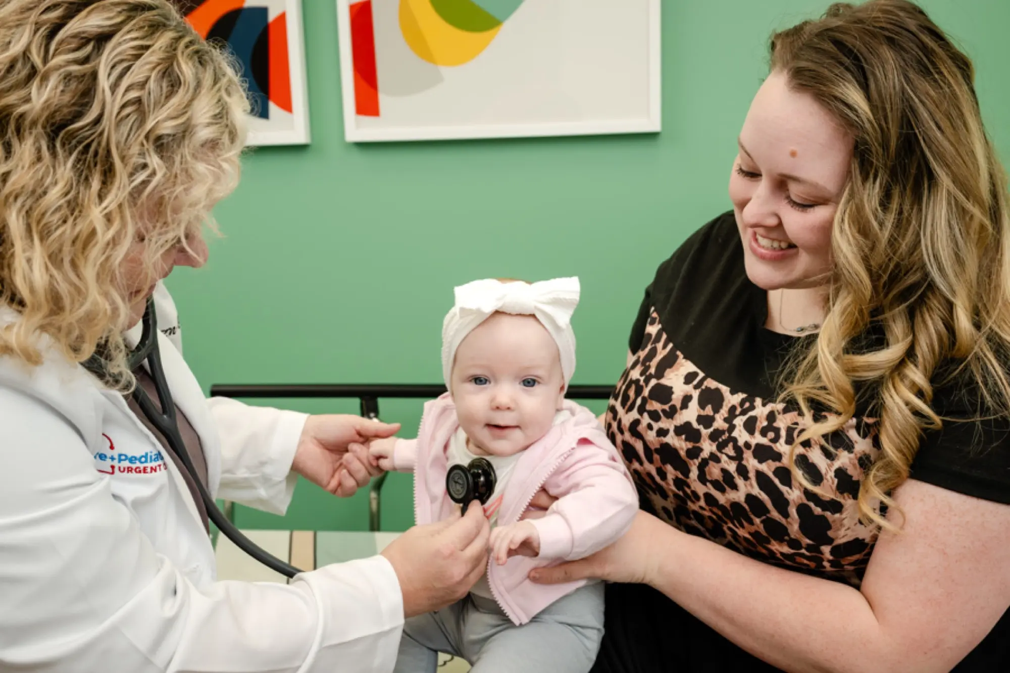 A smiling Care+ provider listening to an infant's heart while the parent holds them close