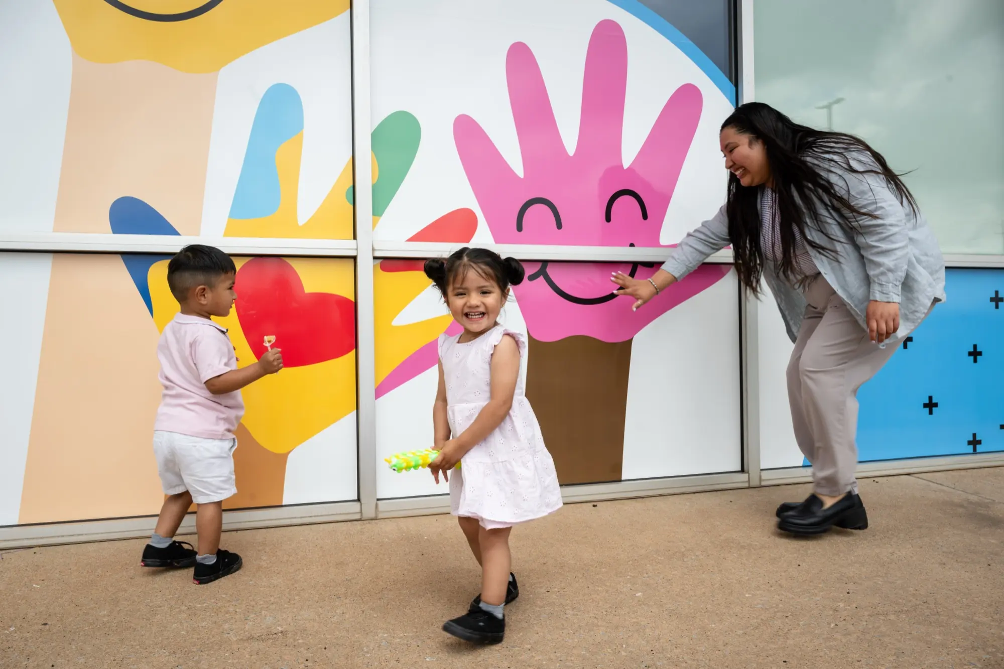 A caregiver playing with two laughing children in front of the colorful hand mural outside the Care+ Edmond clinic
