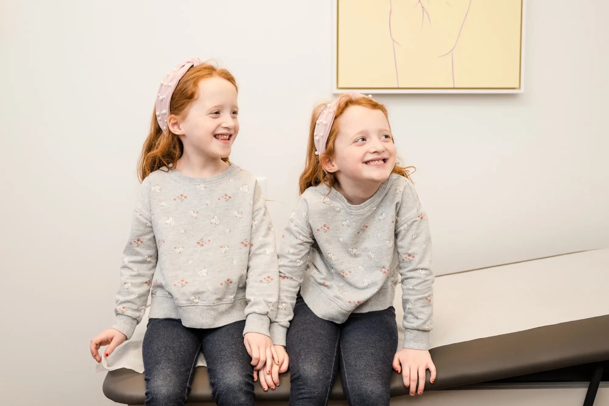 Twin sisters laughing together on the exam table during a visit to Care+ Pediatrics