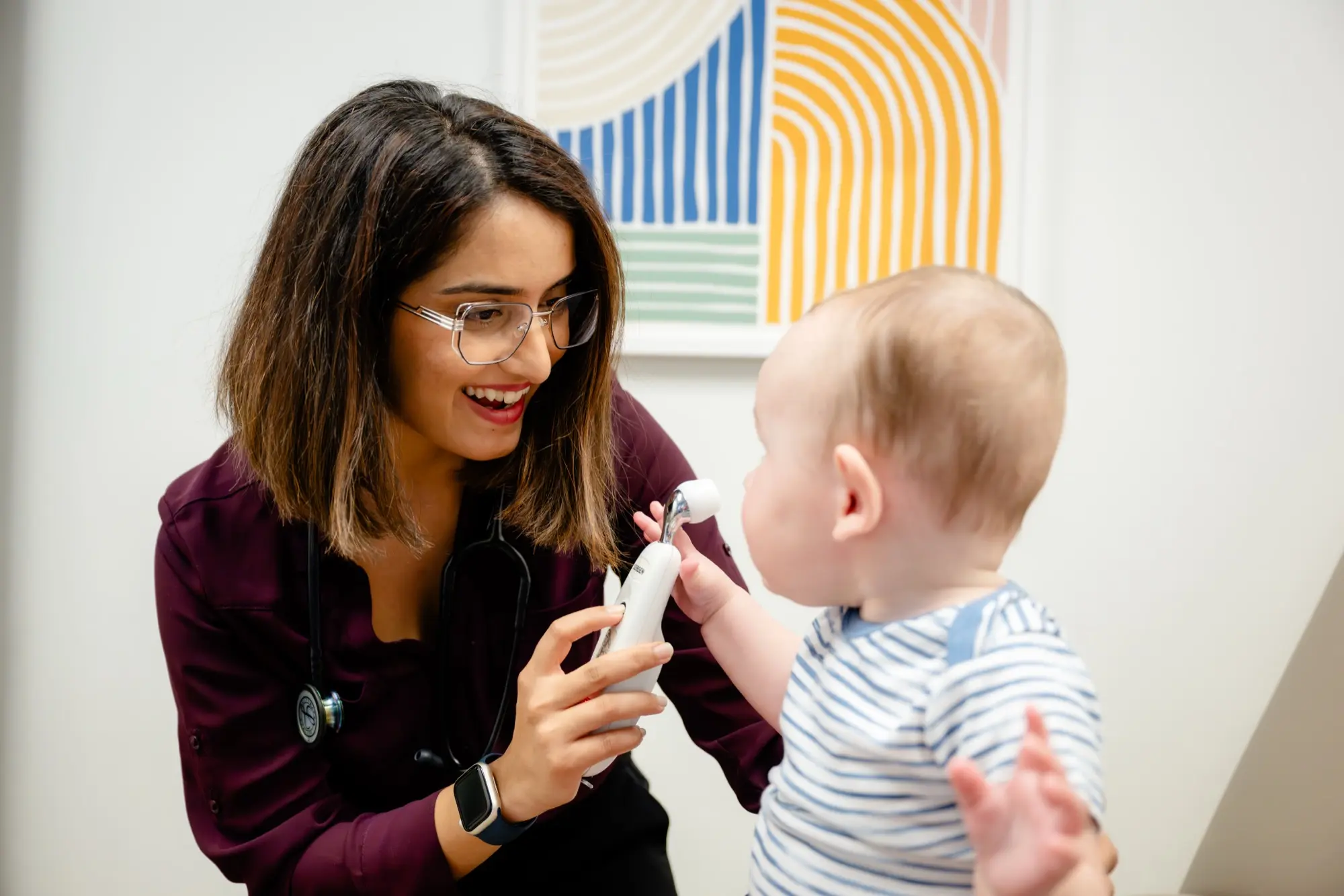 A smiling Care+ provider showing a curious baby the ear thermometer during an exam