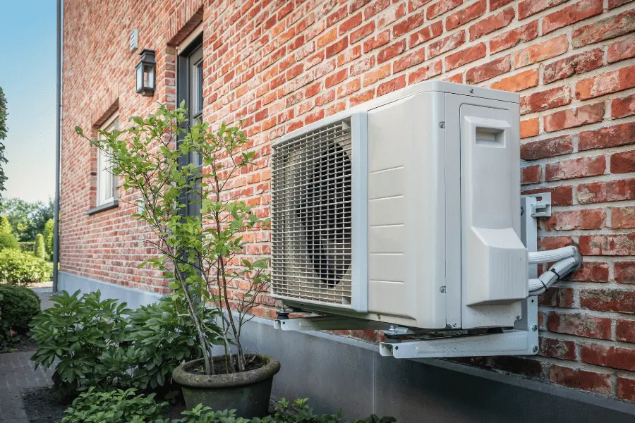 Outdoor air conditioner condenser unit mounted on a brick wall with a metal support bracket, nearby potted plant and shrubs in a sunny yard.