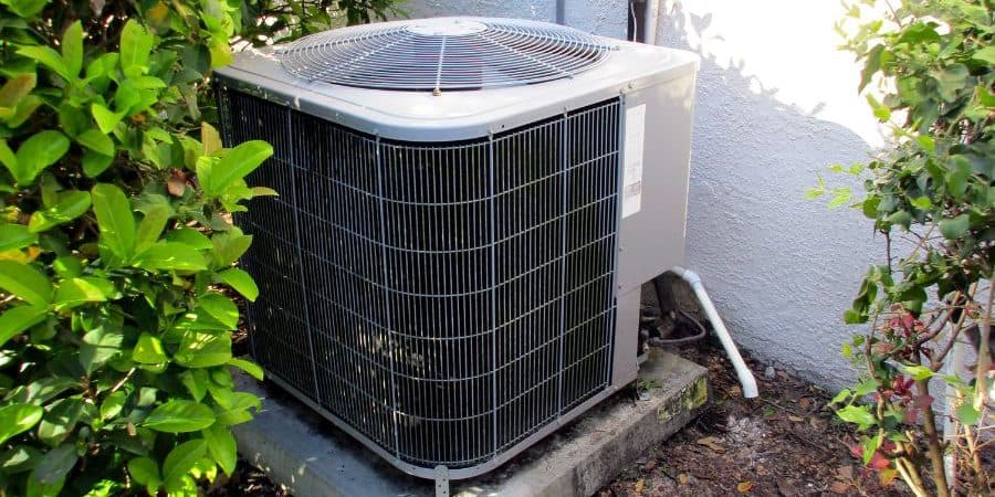 Outdoor air conditioning condenser unit on a concrete pad, surrounded by green shrubs and a white wall nearby.
