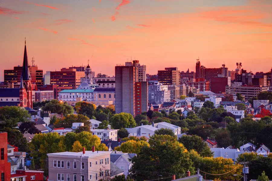 City skyline at sunset with orange-pink sky over a mix of modern and historic buildings and trees in the foreground.